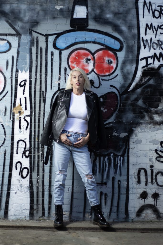 Woman in leather jacket and ripped jeans standing in front of graffiti mural, facing forward with bold expression. Photo credit: Serious Photography.