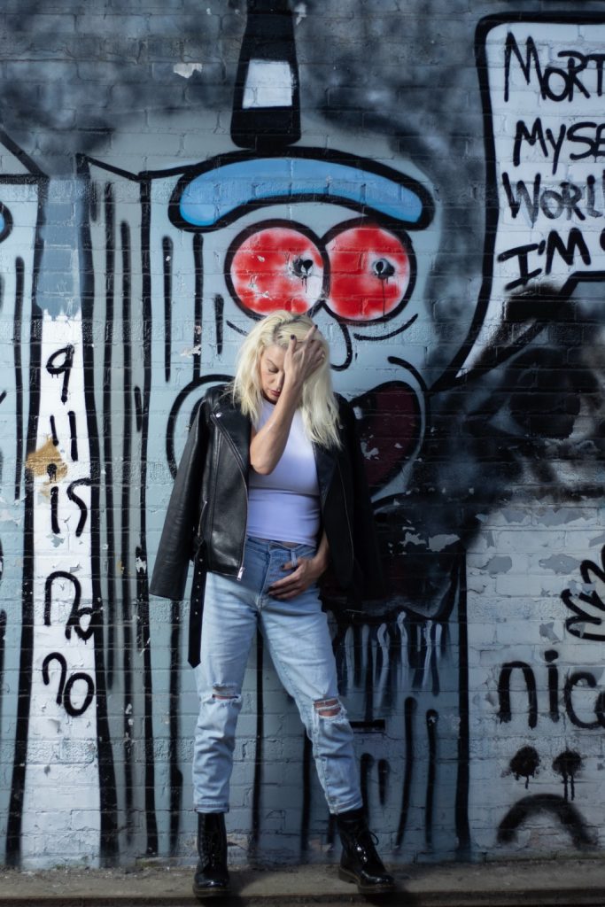 A woman in a leather jacket and ripped jeans, leaning against a graffiti wall, looks down with her hand in her hair, symbolizing resilience and reflection. Photo Credit: Serious Photography.